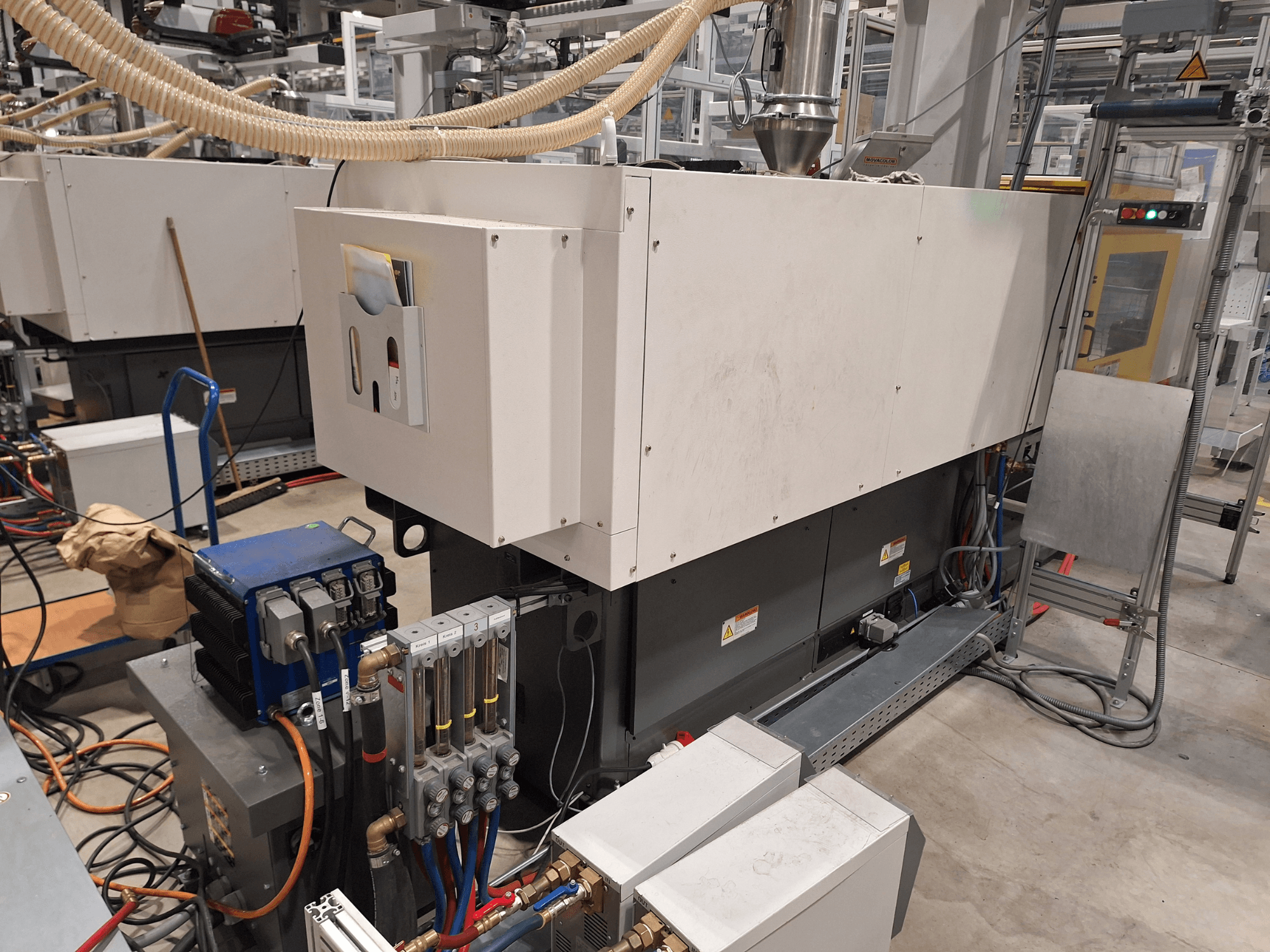 Equipamento industrial numa fábrica; inclui uma máquina de grandes dimensões com painel de controlo, mangueiras e ligações eléctricas, vista lateral.