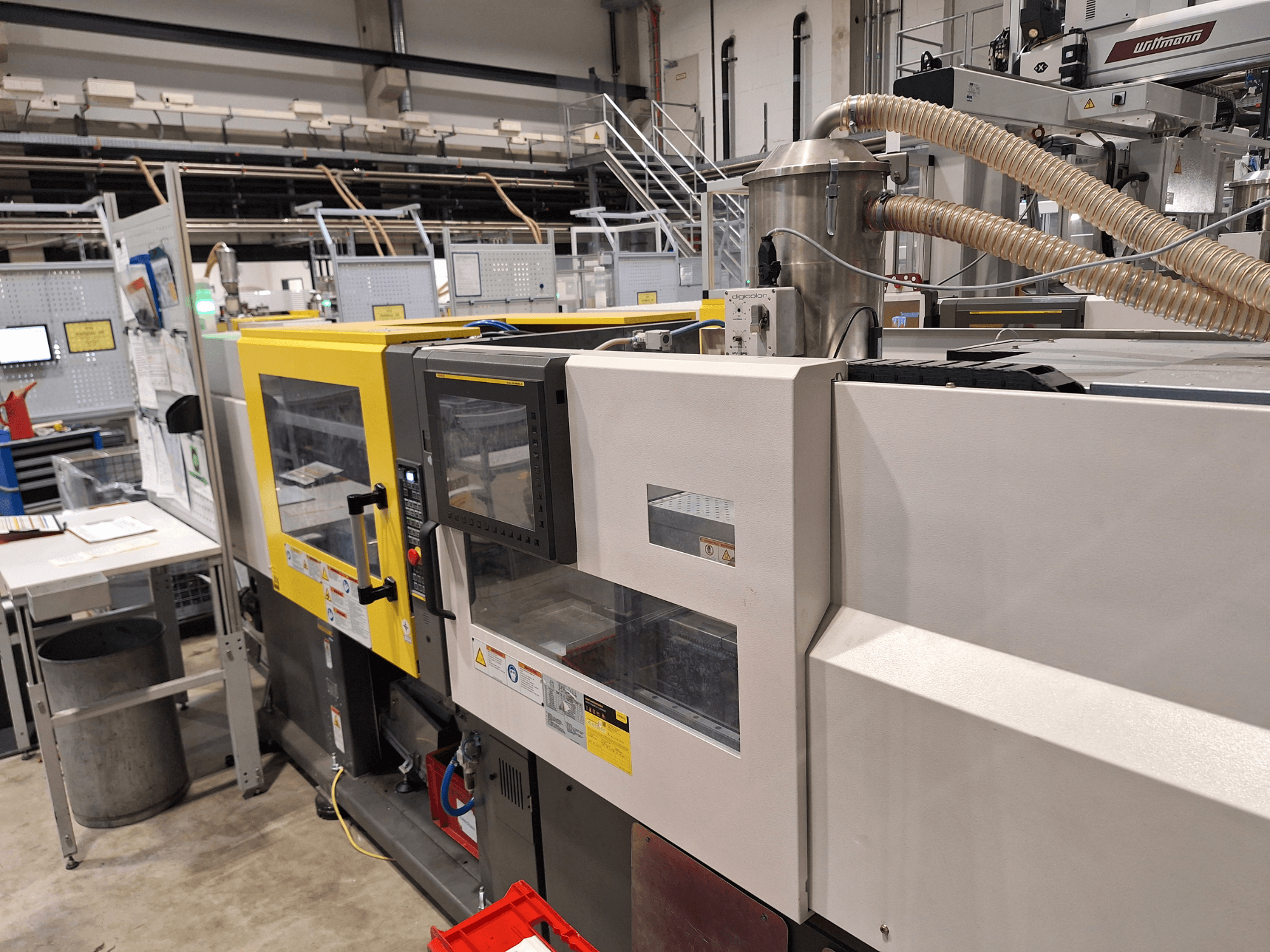 Vista frontal de uma máquina de moldagem por injeção multicolorida, com secções amarelas e cinzentas, com painel de controlo e sistema de escape.
