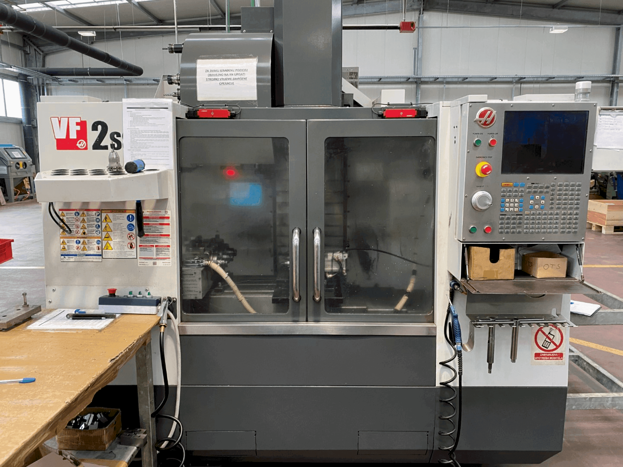 Centro de maquinação Haas VF-2SS em vista frontal, com painel de controlo, mangueira de refrigeração e etiquetas de segurança na lateral.