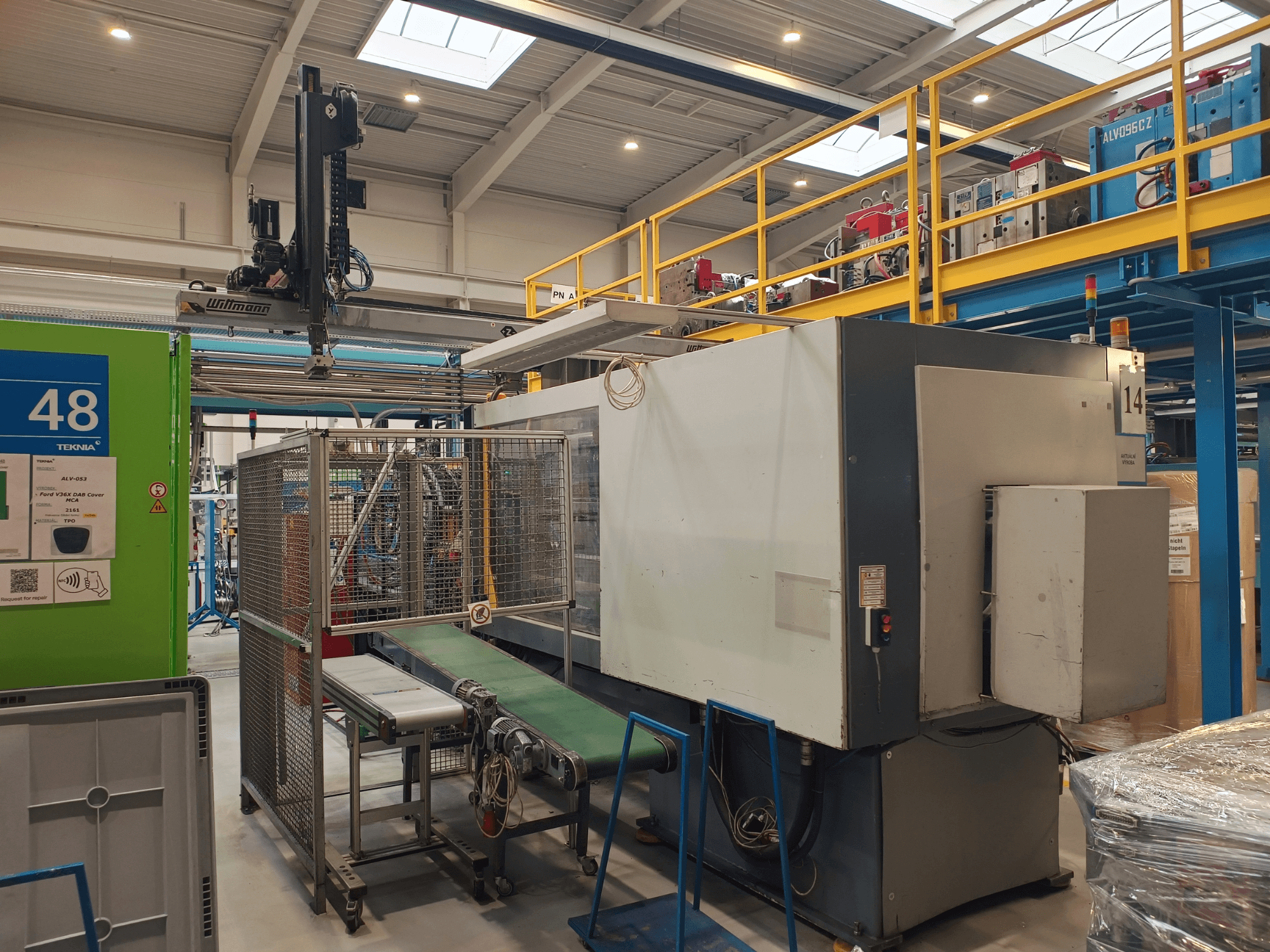 Equipamento de automação industrial com transportador verde, com a etiqueta "48", e ponte rolante; vista lateral da máquina no chão de fábrica.